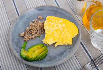 Grey plate with buckwheat, slices of ripe avocado and omelet on the table in the cafeteria