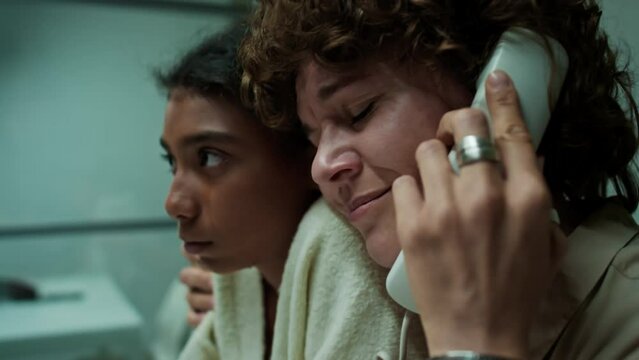 Sad mother hugging her crying teen daughter and speaking on phone with imprisoned husband during visit