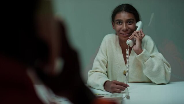 Over the shoulder shot of young African American girl sitting in visiting room, smiling and talking on phone with father serving time in prison