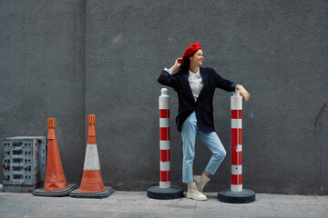 Fashion woman standing leaning against a wall street against a background of the city road works tourist in stylish clothes with red lips and red beret, travel, cinematic color, retro vintage style.