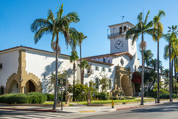 Santa Barbara, CA, USA - November 30, 2023: Santa Barbara County Courthouse, white stone and tower with palm trees and green lawn seen from SW corner against blue sky. Traffic light