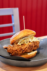 A fried chicken sandwich topped with fresh coleslaw is plated on  a wood table in front of a chair and red wall.