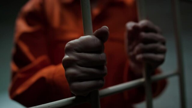 Close up shot of hands of unrecognizable inmate in orange uniform holding bars of prison cell