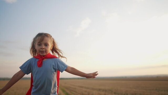 girl with the cloak. concept of a happy childhood and family for a child. a little girl in a superhero costume runs across a field, a red cape and lifestyle a blue T-shirt, sunset on the background