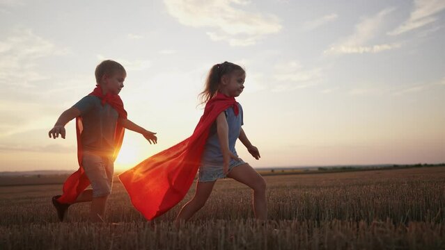 children in superhero costumes. concept of a happy childhood and family for a child. a boy and a girl with red capes and blue T-shirts are running across lifestyle a field, sunset in the background