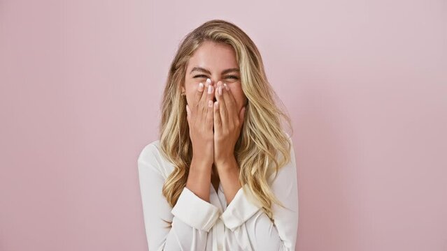 Cheerful blonde gal caught in funny gossip scandal, standing over pink backdrop, wearing a shirt, she covers her laughing mouth, blushing in joyful embarrassment.