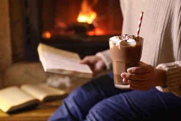Fotobehang Woman with glass of hot cocoa and book near fireplace at home, closeup. Space for text © New Africa