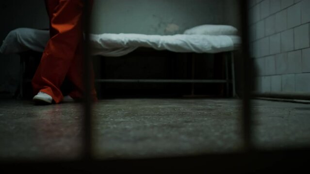 Low section shot through bars of legs of anxious inmate in orange jail uniform pacing around prison cell