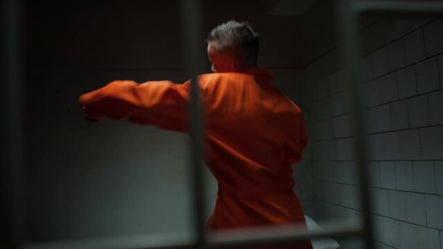 View through bars of brutal female inmate with short hair wearing orange uniform shadowboxing in prison cell to release rage