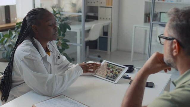 Medium shot of young African American female doctor in white coat sitting at desk in clinic with Caucasian male patient, demonstrating photo on smartphone with healthy foods, giving nutritional advice