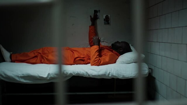 View through bars of African American inmate in orange uniform lying down on bed in prison cell, looking at photos of family on wall and missing them