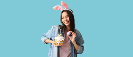 Pretty young woman with bunny ears, Easter cake and egg on light blue background