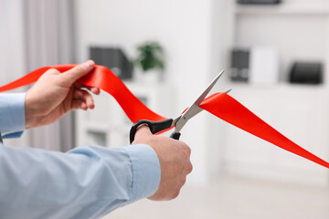 Man cutting red ribbon with scissors indoors, closeup