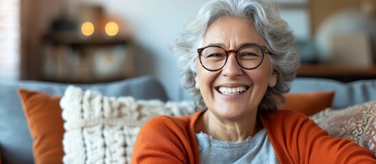 Cheerful older woman in glasses, smiling and laughing on sofa, displaying her healthy teeth while enjoying leisure at home. - obrazy, fototapety, plakaty