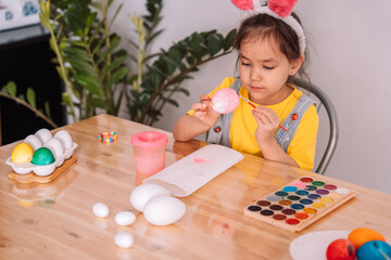 Little girl paints eggs for Easter at a large wooden table.