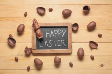 Chalkboard with text HAPPY EASTER, chocolate bunny and eggs on wooden background