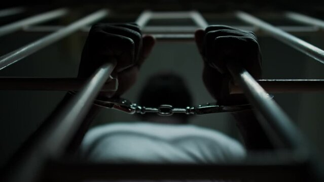 Directly below view of hands of African American inmate in handcuffs holding bars of prison cell