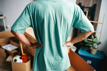 Rear view of young man standing and looking at cardboard boxes in front of him after moving in new...