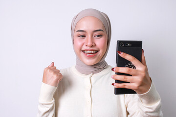 Excited Asian muslim girl with hijab and braces teeth, clenched fist showing victory gesture while looking at mobile phone