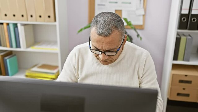 A middle-aged hispanic man in an office setting focused on his computer screen with shelves and plants in the background.