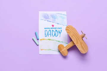 Card for Father's Day with chalks and wooden airplane on lilac background