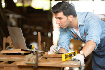 Carpenter working on laptop in his workshop at a woodworking factory - obrazy, fototapety, plakaty