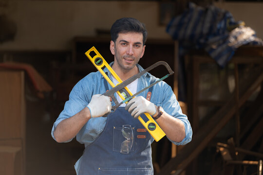 Portrait of a carpenter holding a spirit level and handsaw in his workshop.