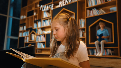 Young smart caucasian girl picking reading a book while sitting at library. Clever child learning,...