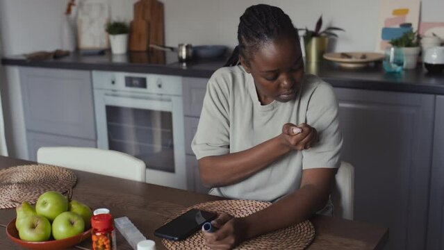 Medium shot of African American woman with braided hair, in casual t-shirt sitting in kitchen, opening insulin pen and injecting in upper arm, medicine containers and fruit on table