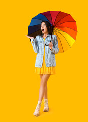 Beautiful shocked young woman with umbrella on yellow background