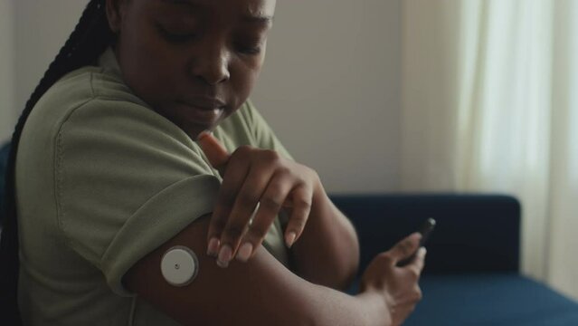 Medium close-up of young African American woman with diabetes sitting on couch in living room, touching blood sugar monitoring device on arm, then checking data on smartphone app