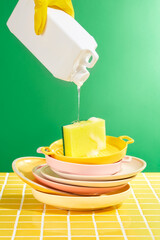 Stacks of ceramic dishes and pans washed on the yellow tile floor. A hand is holding the plastic canister and slowly pouring down the detergent. Professional eco cleaning service supplies.
