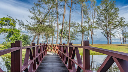 A plank pedestrian bridge with wooden railings runs over a calm river. The ocean is ahead. Green...