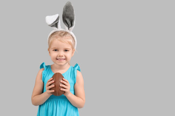 Happy little girl in bunny ears with chocolate egg on grey background. Easter celebration