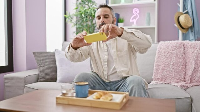 Man photographing breakfast on phone, indoor cozy living room with sofa and decor.