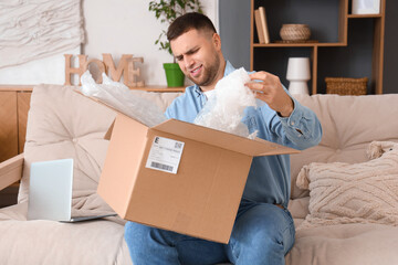 Displeased young man unpacking parcel on sofa at home