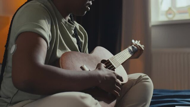 Medium close-up shot of young black woman with diabetes, wearing blood glucose monitor on arm, sitting in bedroom at home, turning on playback on laptop, then playing ukulele guitar and singing