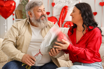 Beautiful mature couple with bouquet of roses sitting on sofa at home. Valentine's Day celebration