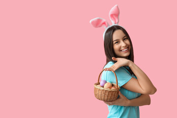 Pretty young woman with bunny ears and basket of Easter eggs on pink background