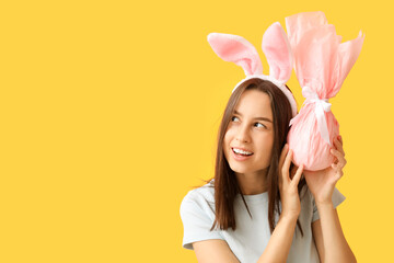 Pretty young woman with bunny ears and Easter gift on yellow background