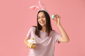 Pretty young woman with bunny ears, Easter cake and egg on pink background