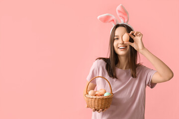 Pretty young woman with bunny ears and basket of Easter eggs on pink background