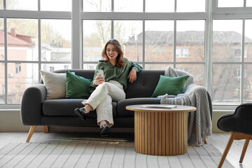Young woman using mobile phone on sofa near window in living room