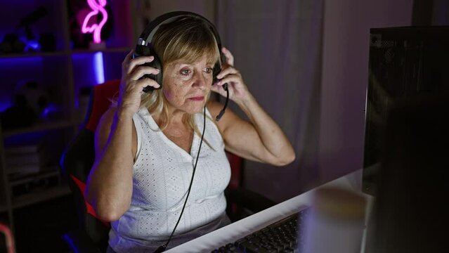 Smiling middle age blonde bombshell streaming, wired in play mode, gaming room vibing. this beautiful woman, wearing headphones, takes on the digital world night after night.