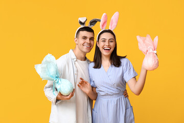 Happy young couple with Easter bunny ears and gifts on yellow background