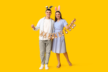Happy young couple with Easter bunny ears and garland on yellow background