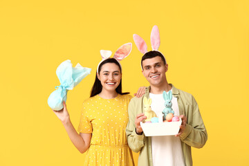 Happy young couple with Easter bunny ears, basket and gift on yellow background