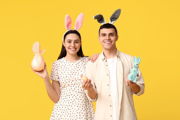 Happy young couple with Easter bunny ears, gift, toy rabbit and egg on yellow background