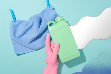 A cloth towel held in place by two clothespins, a hand in a pink rubber glove, acrylic sheet and a bottle of detergent on a blue background. Blank label for branding.