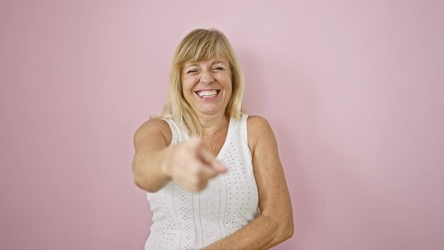 Confident middle age blonde woman bursting with laughter, pointing joyfully at the camera over isolated pink background.